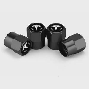 May include: Four black tire valve stem caps with a white Tesla logo. The caps are hexagonal and have a glossy finish. The Tesla logo is a stylized letter "T" in white.