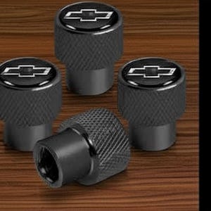 May include: Set of four black tire valve stem caps with a textured finish. Each cap features a silver Chevrolet bow tie logo on a black background. The caps are displayed on a wooden surface.