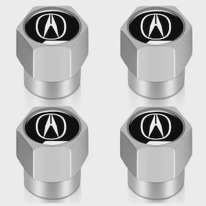 May include: Set of four silver-colored tire valve stem caps. Each cap features a black oval with a white Acura logo. The caps have a hexagonal shape and a metallic finish, suitable for car enthusiasts.