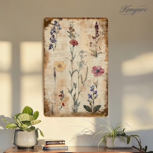 Può includere: Un cartello metallico rettangolare con illustrazioni botaniche di vari fiori nei toni del viola, rosso, giallo e rosa. Lo sfondo ha un aspetto vintage e invecchiato con testo simile a una sceneggiatura. Il cartello ha un bordo marrone e la parola "Kanyawe" nell'angolo in alto a destra.