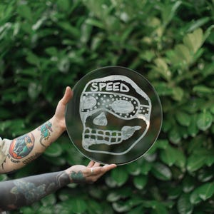 Może przedstawiać: Przezroczysty, okrągły szklany talerz z wytrawionym wzorem czaszki. Słowo "SPEED" znajduje się nad czaszką. Talerz jest trzymany na tle zielonych liści.