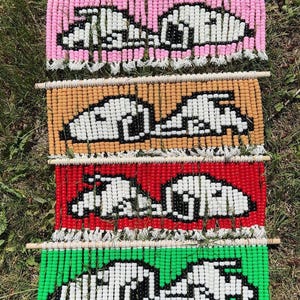 May include: Four rectangular wall hangings made of colorful plastic beads strung on white cord and suspended from wooden dowels. Each piece features a white and black beaded graphic of a dog lying down, set against backgrounds of pink, tan, red, and green.