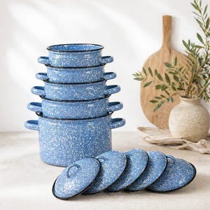 Puede incluir: Un juego de seis ollas de esmalte azul con tapas. Las ollas están apiladas, con las tapas dispuestas al frente. Tienen un patrón blanco moteado y ribete negro. Una tabla de cortar de madera y un jarrón con vegetación están en el fondo.