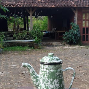 Könnte beinhalten: Eine Vintage-Teekanne aus Emaille mit einem grün-weißen Sprenkelmuster. Die Teekanne hat einen Ausguss, einen Griff und einen Deckel und steht auf einem Holztisch. Im Hintergrund ist ein traditionelles indonesisches Haus zu sehen.