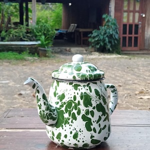 May include: A white and green enamel teapot with a speckled pattern. The teapot has a lid, spout, and handle. It is sitting on a wooden table outdoors.