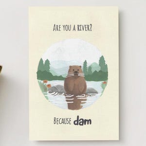 Könnte beinhalten: Grußkarte mit einer Biber-Illustration in einem Kreis mit Bäumen und Wasser. Der Text lautet "Are you a river? Because dam". Die Karte liegt auf einer weißen Oberfläche mit einem Tablett mit Büroklammern und einer Tasse Kaffee.
