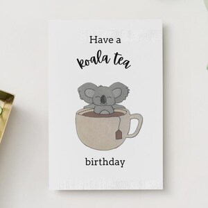 Könnte beinhalten: Eine weiße Karte mit dem Text "Have a koala tea birthday" und einer Illustration eines Koalas in einer Teetasse. Ein goldfarbenes Tablett mit grünen Büroklammern, eine Tasse Kaffee und ein Keks sind ebenfalls zu sehen.