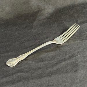 May include: A silver fork with four tines and a decorative handle. The handle features an ornate floral design. The fork is made of a shiny, metallic material and is set against a dark background.