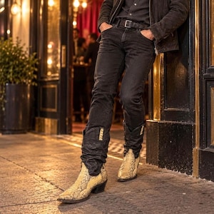 May include: A pair of black denim jeans with a leather belt and a dark jacket, paired with pointed-toe cowboy boots featuring a textured, cream and tan snakeskin pattern. The boots have a stacked heel and a distinct, scrunched leather shaft design.