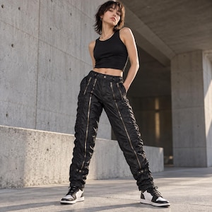May include: A black sleeveless tank top paired with black trousers featuring prominent vertical zippers running down the front of each leg, finished with black and white athletic sneakers. The trousers have a textured, slightly ruched appearance.