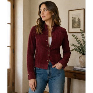 May include: A burgundy suede jacket with a stand-up collar and long sleeves. The jacket features gold-colored buttons down the front and on the cuffs. The model is wearing blue jeans.