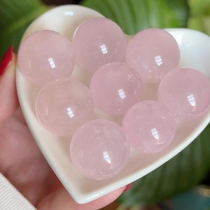 May include: Seven smooth, polished rose quartz spheres arranged in a white, heart-shaped ceramic dish. Each sphere exhibits a translucent, pale pink hue with natural internal inclusions and a reflective, glossy surface finish throughout.