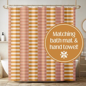 Puede incluir: Una cortina de ducha con un patrón repetitivo de rayas horizontales en tonos naranja quemado, crema y líneas finas de color rojo oscuro. Una superposición circular contiene el texto Matching bath mat & hand towel! con un pequeño icono floral.