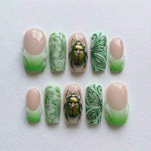 May include: A set of ten artificial nails with a variety of designs. The nails feature a light pink base with green and white accents. Designs include a lizard, a beetle, and a butterfly wing.