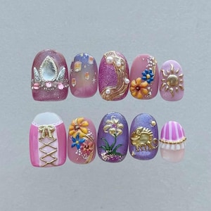 May include: A set of ten artificial nails with a princess theme. The nails feature various designs, including a crown, floating lanterns, flowers, and a sun, in shades of pink, purple, and gold. Each nail has a unique design.