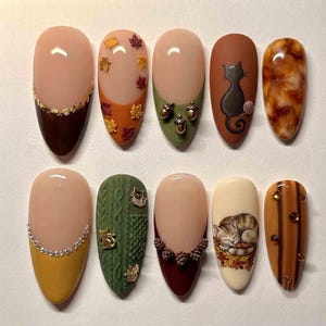 May include: A set of ten almond-shaped press-on nails featuring autumn-themed designs. The collection includes French tips with gold leaf, maple leaf accents, 3D acorns, a black cat silhouette, marble patterns, rhinestones, cable knit textures, pinecones, and a sleeping cat illustration.