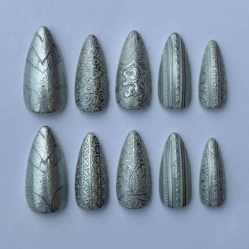May include: A collection of ten silver-toned, almond-shaped false nails. Each nail is decorated with detailed embossed patterns, such as geometric shapes, floral designs, and a heraldic lion. The nails are displayed in two rows of five.