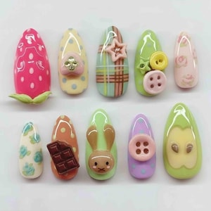 May include: A collection of ten artificial nails with various designs. The nails feature a strawberry, polka dots, plaid, buttons, flowers, a chocolate bar, a bunny, and an apple. The nails are painted in a variety of colors, including pink, yellow, green, and purple.