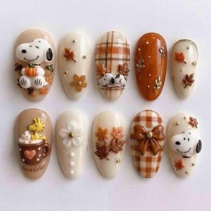 May include: A set of ten press-on nails featuring autumn themes, including Snoopy and Woodstock characters, brown and white plaid patterns, 3D sculpted leaves, a brown bow, and a mug with the text Good Grief, accented with small gold beads.