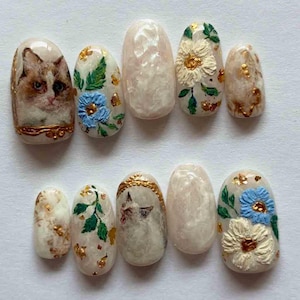 May include: Set of ten artificial nails with various designs. Some nails feature a cat's face, others have floral patterns with blue and white flowers, green leaves, and gold accents. The nails have a glossy finish and are oval-shaped.