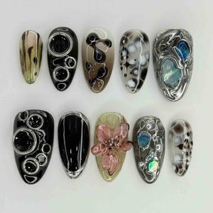 May include: A collection of ten oval-shaped artificial nails with various designs. The designs include abstract patterns, bubble accents, and a pink floral embellishment. Colors range from black and white to iridescent and neutral tones.