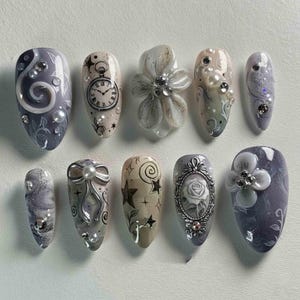 May include: A set of ten artificial nails in various designs. The nails feature a mix of gray, white, and beige colors with embellishments like pearls, rhinestones, and 3D floral accents. Designs include swirls, clocks, bows, stars, and a rose.