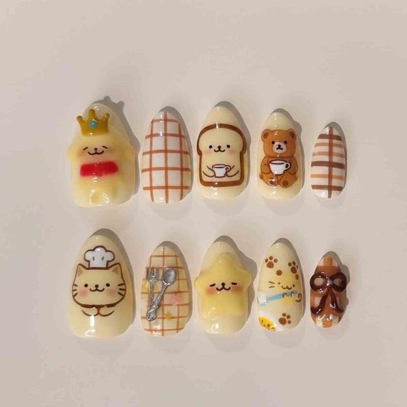 K&ouml;nnte beinhalten: Ein Set aus zehn k&uuml;nstlichen N&auml;geln mit verschiedenen Designs. Die N&auml;gel zeigen Cartoon-Charaktere, Muster zum Thema Essen und ein kariertes Muster in Braun-, Gelb- und Wei&szlig;t&ouml;nen. Einige N&auml;gel haben ein gl&auml;nzendes Finish, andere sind matt.