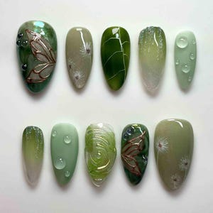 May include: A set of ten artificial nails in various shades of green. Designs include butterfly wings, leaf patterns, and water droplet accents. Some nails feature a gradient effect, while others have delicate floral details.