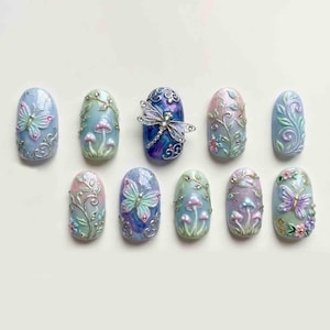 May include: A collection of ten intricately designed nail art pieces. Each nail features a different design, including butterflies, mushrooms, and floral patterns, in pastel colors with silver accents. The nails are oval-shaped and arranged on a white surface.