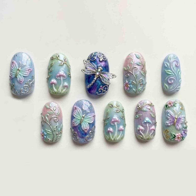May include: A collection of ten intricately designed nail art pieces. Each nail features a different design, including butterflies, mushrooms, and floral patterns, in pastel colors with silver accents. The nails are oval-shaped and arranged on a white surface.