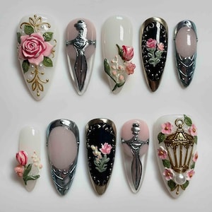 May include: A collection of ten almond-shaped artificial nails with various designs. The designs include 3D roses, a sword, a lantern, and floral accents in pink, gold, silver, and black. The nails are set against a white background.