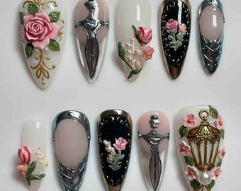 May include: A collection of ten almond-shaped artificial nails with various designs. The designs include 3D roses, a sword, a lantern, and floral accents in pink, gold, silver, and black. The nails are set against a white background.