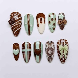 May include: A set of ten almond-shaped artificial nails with various chocolate-themed designs. The designs include chocolate drips, a chocolate bar, hearts, stars, cookies, and bows. Colors include brown, white, and mint green.