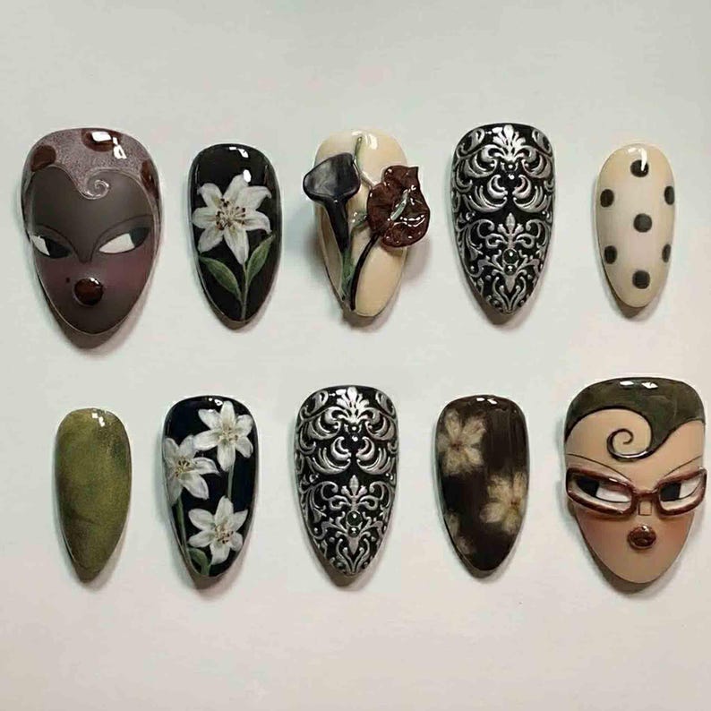 Puede incluir: Una colecci&oacute;n de diez u&ntilde;as artificiales con varios dise&ntilde;os. Los dise&ntilde;os incluyen patrones florales, caras abstractas y filigranas ornamentadas. Los colores incluyen negro, blanco, beige, marr&oacute;n y verde oliva. Las u&ntilde;as son de forma ovalada y parecen estar hechas profesionalmente.