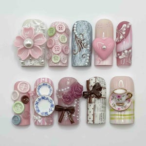 May include: A set of ten artificial nails with diverse designs. The designs include a pink flower with a pearl centre, buttons, lace, a heart, and miniature plates. Other nails feature roses, bows, and a teacup. The nails are in shades of pink, white, and beige.