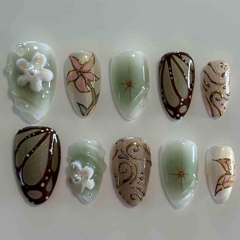 Peut inclure: Dix faux ongles avec divers motifs. Les ongles pr&eacute;sentent des motifs floraux, papillons et abstraits dans des tons de vert, marron, or et blanc. Certains ongles ont des accents floraux en 3D.