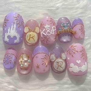 May include: A set of ten artificial nails with a Disney theme. The nails feature a variety of designs, including a castle, fireworks, Mickey Mouse heads, initials, and a crown. Colors include lavender, pink, and glitter accents.