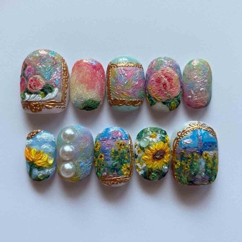Op de afbeelding: Een set van tien kunstnagels met ingewikkelde ontwerpen. Elke nagel heeft een uniek, kleurrijk ontwerp, waaronder bloempatronen, parels en landschapssc&egrave;nes. De nagels zijn ovaal van vorm en hebben een glanzende afwerking, wat gedetailleerde kunstzinnigheid laat zien.