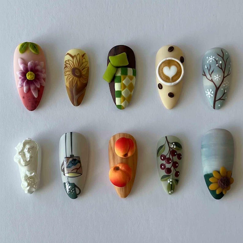 May include: A collection of ten almond-shaped nail designs. The designs include a pink and purple flower, a sunflower, a green scarf, a coffee cup with a heart, a winter tree, a white sculpture, a lamp, two peaches, cherries, and a sunflower.
