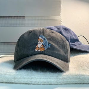 May include: A gray baseball cap with a curved brim. The cap features an embroidered design of a cat wearing a shark costume. A second blue cap is visible in the background.