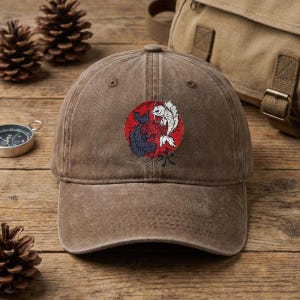 May include: Brown baseball cap featuring a red circle with a black and white koi fish design. The cap has a curved brim and an adjustable strap. The Japanese character for water is below the fish.
