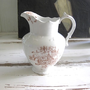 May include: A white ceramic pitcher featuring a brown floral transferware pattern. The vessel has a scalloped rim, a curved handle, and a rounded body. The brown design depicts detailed clusters of chrysanthemums and foliage on the front surface.