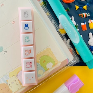 Peut inclure: Un agenda avec un calendrier, des autocollants et des articles de papeterie. L'agenda est ouvert sur une page avec un calendrier. Un surligneur bleu clair et une gomme rose sont sur la page. Un tampon rectangulaire rose avec des motifs de lapin est également présent.