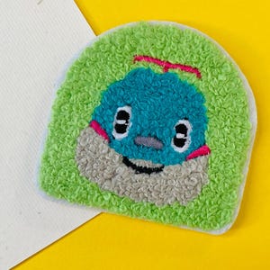 Puede incluir: Un parche bordado a mano con un personaje de dibujos animados. El parche tiene una cara azul, una boca gris y un fondo verde texturizado. El personaje tiene ojos negros y detalles rosas. El parche está sobre un fondo blanco y amarillo.