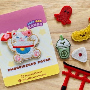 May include: Embroidered patches with Japanese themes. Designs include a waving cat, a red torii gate, a red octopus, a tempura shrimp, a green tea drink, and rice balls. The patches are on a wooden surface.