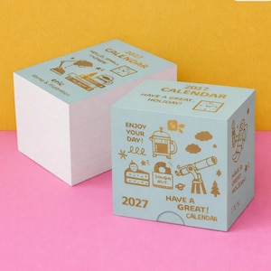 May include: Two stacks of light blue paper calendars for 2027. The top sheets feature gold-colored illustrations including a telescope, a French press, a slice of cake, a donut, and an astronaut, with text reading 2027 CALENDAR HAVE A GREAT HOLIDAY.