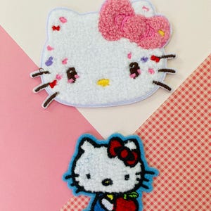May include: Two embroidered patches featuring a cartoon character. One has a pink bow and colourful accents. The other has a red bow and holds a red apple. Both have a white background and black outlines.