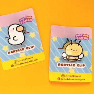 Puede incluir: Dos clips acrílicos sobre tarjetas con fondo amarillo y rosa. Un clip es un pato blanco con pico marrón y brazos de palo. El otro es una abeja amarilla y negra. Las tarjetas tienen el texto "JUST ADD KAWAII" y "ACRYLIC CLIP".