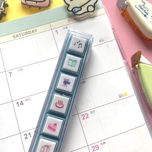 Puede incluir: Un juego de sellos de plástico transparente con seis sellos cuadrados con iconos como notas musicales, un libro, una cámara de cine, una fuente termal, un pastel y una maleta. Los sellos están en un calendario.