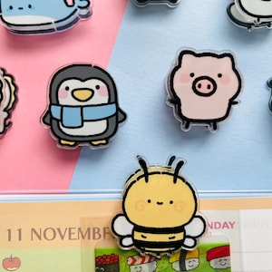 May include: Acrylic magnets with cartoon animal designs, such as a penguin with a blue scarf, a pig, a bee, and a whale. The magnets are displayed against a pink and blue backdrop, with a calendar page visible.